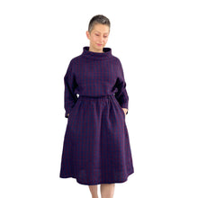 Load image into Gallery viewer, Edith sewing pattern – mid-length linen dress with elasticated waist, long wide sleeves, and funnel neck, front view in plum and navy checkered fabric, model with both hands in pockets