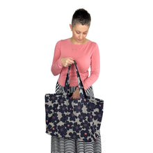 Load image into Gallery viewer, Kulmi Bag sewing pattern by Dhurata Davies – held in right hand with left hand reaching into front pocket, made in light grey canvas with large dark flowers.