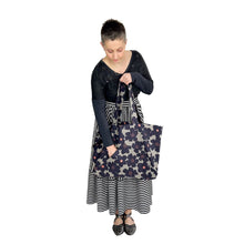 Load image into Gallery viewer, Kulmi Bag sewing pattern by Dhurata Davies – held in left hand with right hand reaching into front triangular pocket, made in light grey canvas with dark flowers and pink centres.