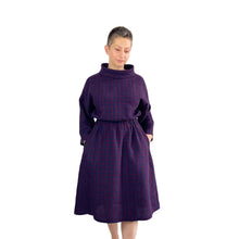Load image into Gallery viewer, Edith sewing pattern – mid-length linen dress with elasticated waist, long wide sleeves, and funnel neck, front view in plum and navy checkered fabric, model with both hands in pockets