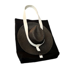 Load image into Gallery viewer, A black fabric Hattie tote bag with white shoulder straps, shown on a white background with a dark woven hat secured over the front by the strap.