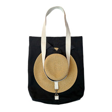 Load image into Gallery viewer, A black canvas tote bag with cream-colored shoulder straps, featuring a large circular straw hat hanging from the front and secured with a metal clasp.