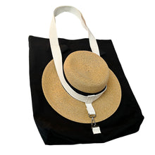 Load image into Gallery viewer, A black fabric tote bag with a wide white shoulder strap, shown holding a woven straw sun hat within.