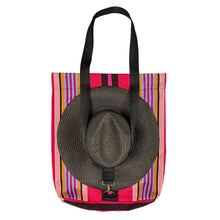 Load image into Gallery viewer, A tote bag with a vibrant pink, orange, and purple striped pattern, featuring black shoulder straps and a large woven straw hat secured over the front by the strap.