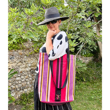 Load image into Gallery viewer, A woman wearing a black wide-brim hat and sunglasses stands outdoors in front of green foliage, holding a large tote bag. The bag has vertical stripes in hot pink, purple, yellow, and black with a black handle.