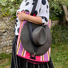 Load image into Gallery viewer, A person outdoors wearing a white blouse with black polka dots and a black skirt, carrying a colorful striped tote bag with a large black woven sun hat strapped over the bag.