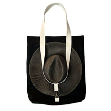 Load image into Gallery viewer, A black canvas tote bag with cream-colored shoulder straps, featuring a large woven straw hat secured over the front by the strap.