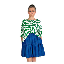 Load image into Gallery viewer, Woman wearing the Rea Top sewing pattern in green and white print, styled with a blue mini skirt