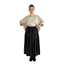 Load image into Gallery viewer, Woman wearing the Tia Skirt sewing pattern in midi length in black fabric with beige stripes, holding the waistband to show the elasticated detail, styled with a beige top