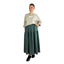 Load image into Gallery viewer, Woman wearing the Tia Skirt sewing pattern in teal in midi length, styled with a natural linen top, hands in pockets