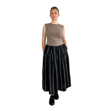 Load image into Gallery viewer, Woman wearing the Tia Skirt sewing pattern in midi length in black fabric with beige stripes, styled with a sleeveless brown top, hands in pockets