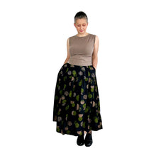 Load image into Gallery viewer, Woman wearing the Tia Skirt sewing pattern in midi length, in black fabric with green and brown dots, worn with a sleeveless brown top, hands in pockets.