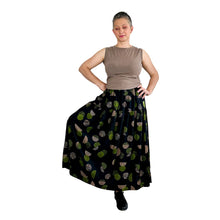 Load image into Gallery viewer, Woman wearing the Tia Skirt sewing pattern in midi length, in black fabric with green and brown dots, worn with a sleeveless brown top, hands on hips.