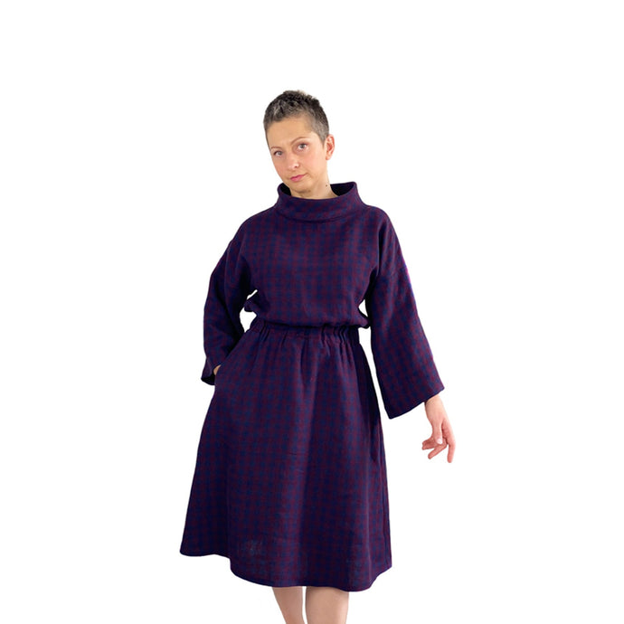 Edith sewing pattern – mid-length linen dress with elasticated waist, long wide sleeves, and funnel neck, front view in plum and navy checkered fabric, model with right hand in pocket