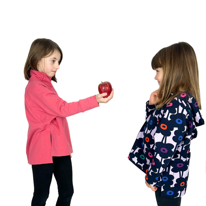 wren pullover hoodie and sweatshirt sewing pattern by dhurata davies, modelled in pink and multicolour floral fabric. the model in pink is holding an apple in her hand